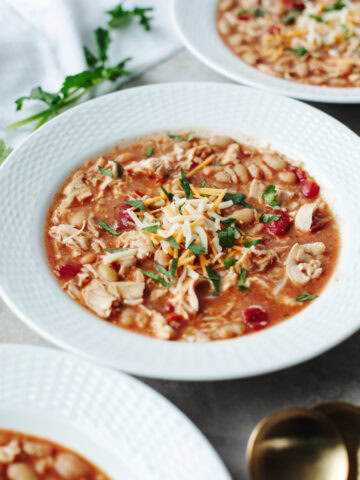 A white bowl filled with hearty chili topped with cilantro, shredded cheese.