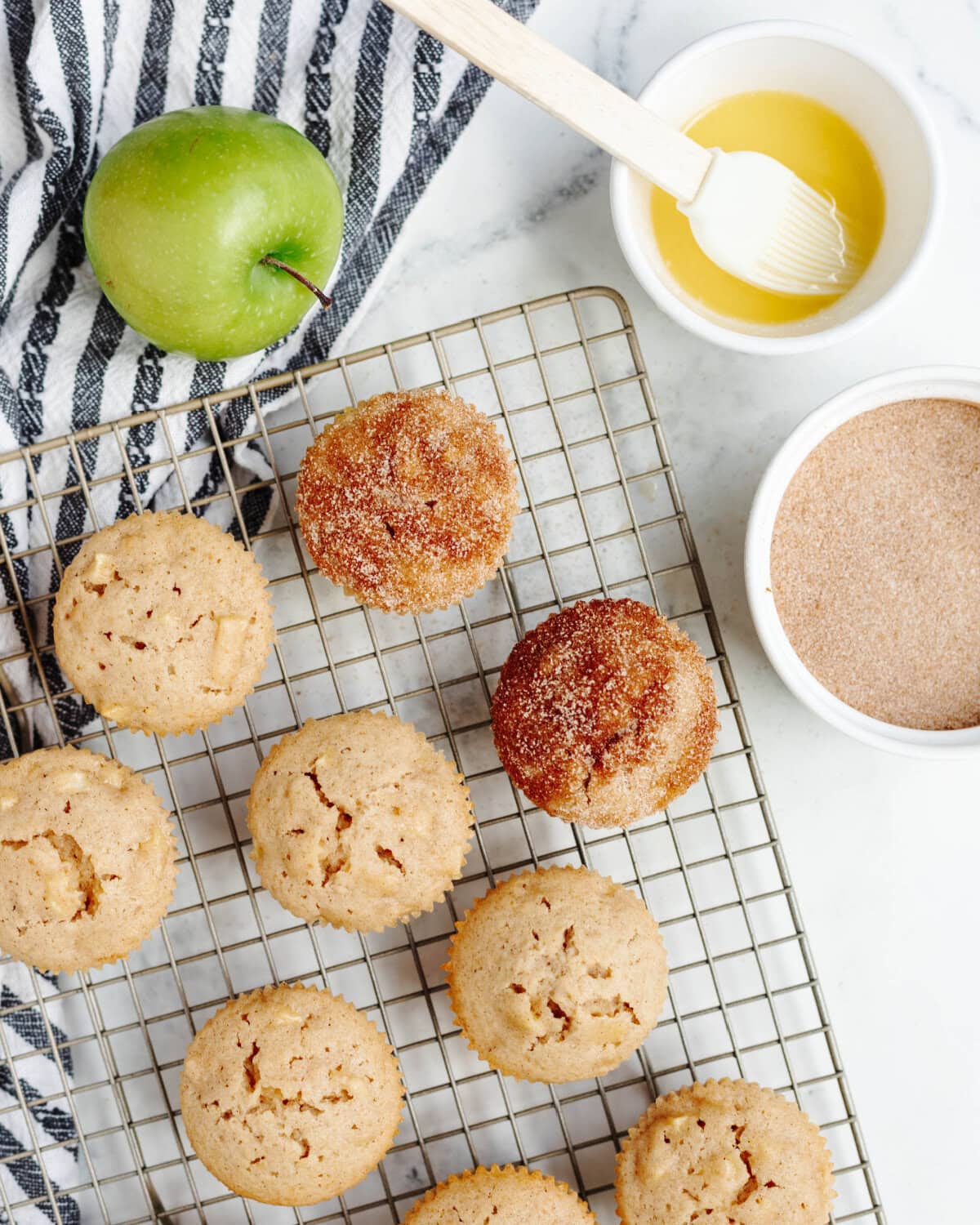 A cooling rack lined with apple muffins getting a cinnamon sugar coating brushed on.