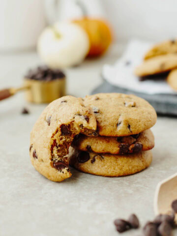 A stack of pumpkin chocolate chip cookies with a bite taken out to show the ooey gooey soft center.
