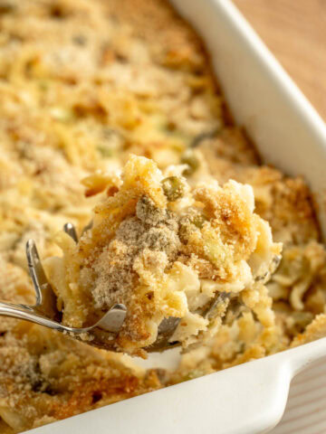 A spoon scooping up a serving of tuna casserole from a white baking pan.