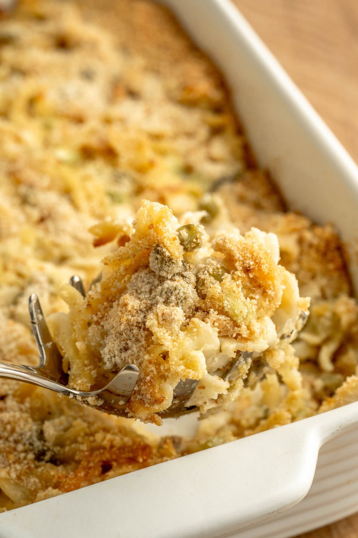 A spoon scooping up a serving of tuna casserole from a white baking pan.